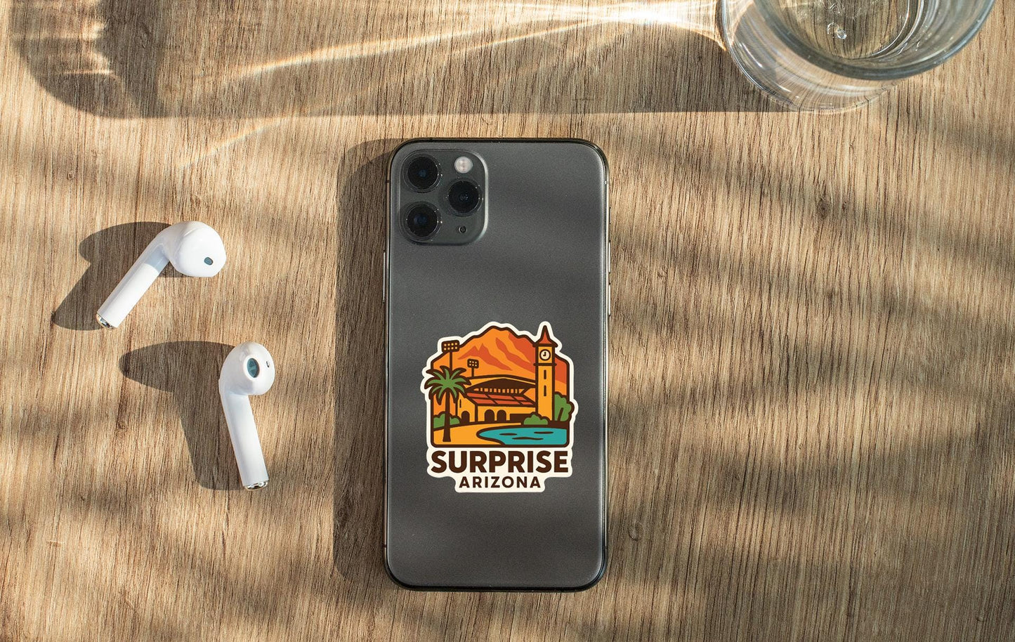 a smartphone with a sticker of Surprise Arizona on its back, along with a pair of white earbuds and a glass of water on a wooden surface.