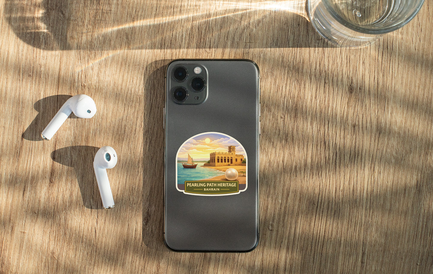 A smartphone with a sticker of a beach and sailboat on the back, along with a pair of white earbuds and a glass of water on a wooden surface.
