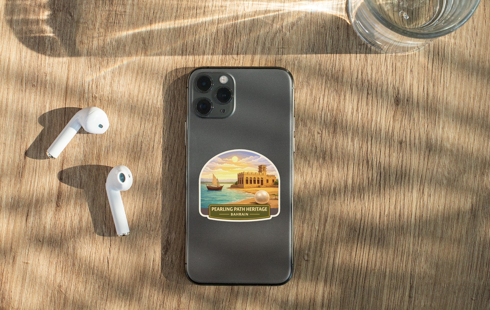 A smartphone with a sticker of a beach and sailboat on the back, along with a pair of white earbuds and a glass of water on a wooden surface.