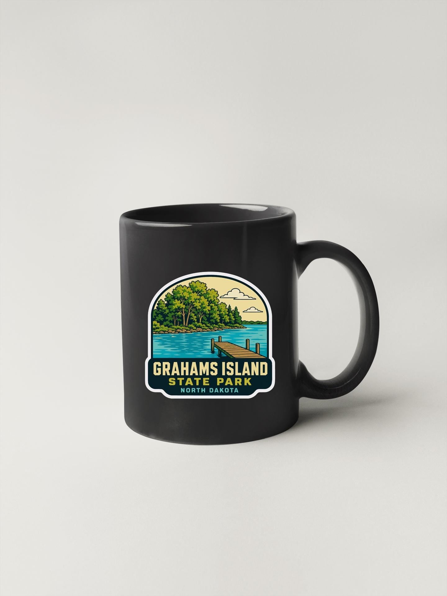 A black coffee mug with a graphic design depicting a scenic view of Grahams Island State Park, featuring a lake, dock, and trees.