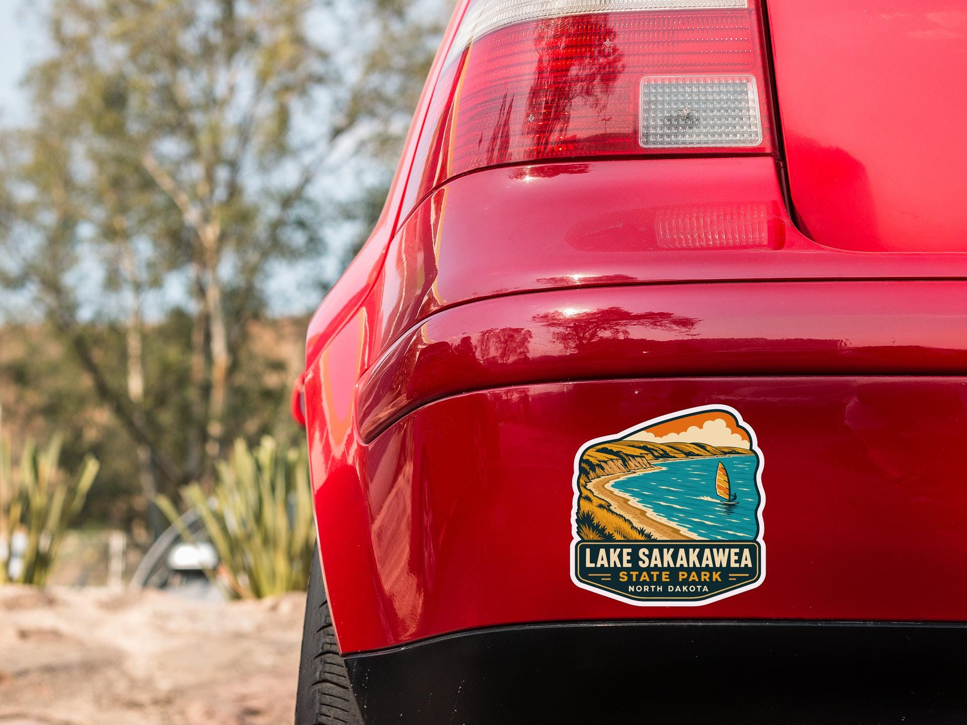 the rear end of a red car with a sticker on the back, featuring a scenic landscape of Lake Sakakawea State Park in North Dakota.