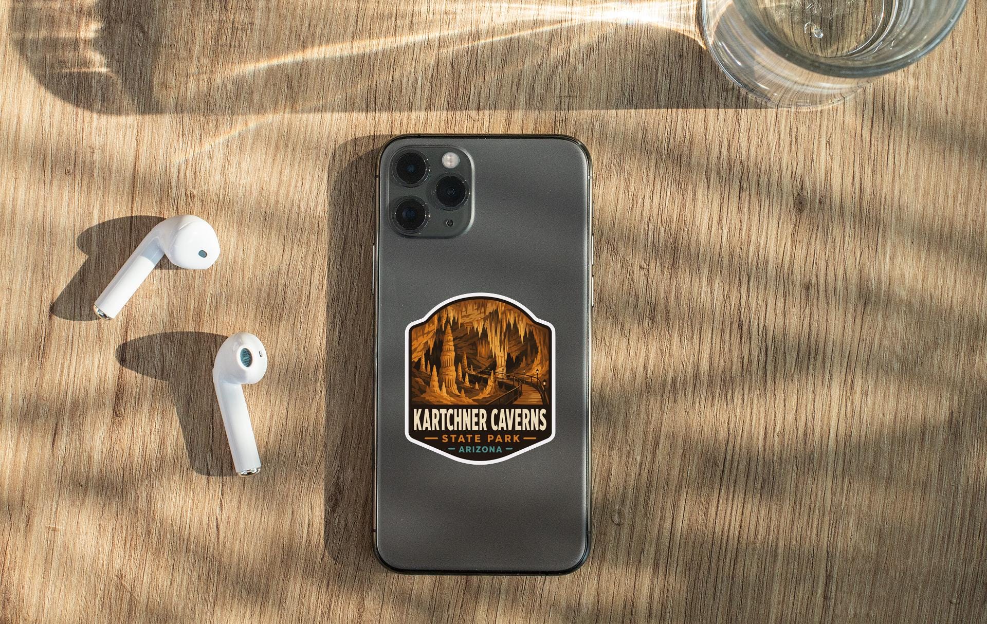 a smartphone with a sticker on it, along with a pair of white earbuds and a glass of water on a wooden surface.