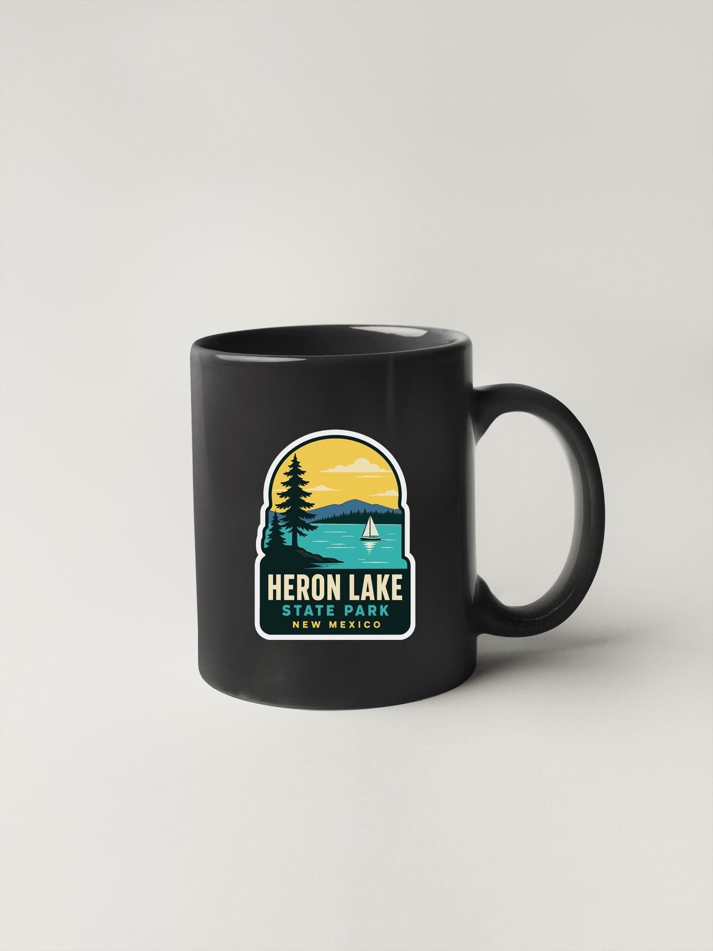 A black coffee mug with a graphic design depicting a scenic lake and mountains, along with the text "HERON LAKE STATE PARK NEW MEXICO".