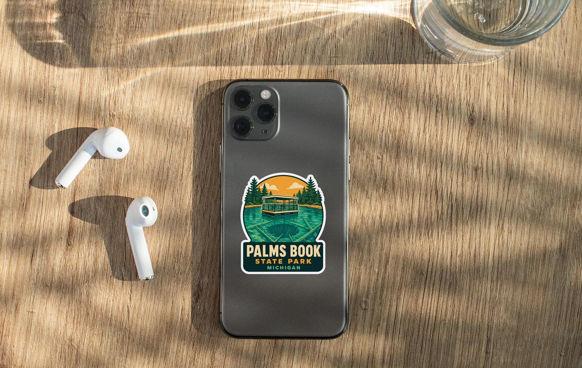 a smartphone with a sticker on it, placed on a wooden surface alongside a pair of white earbuds and a glass of water.
