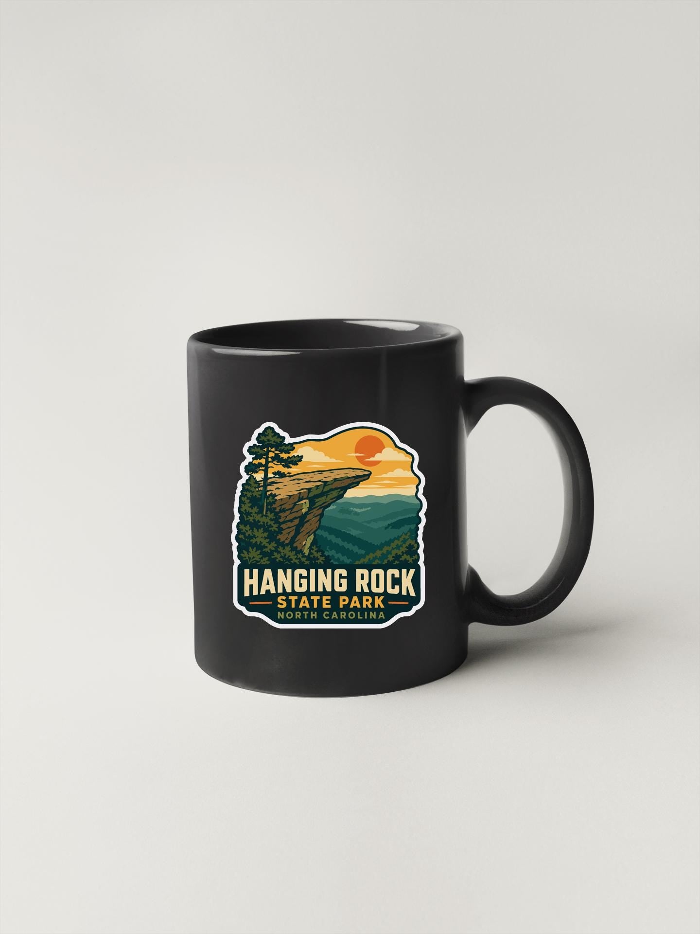 A black coffee mug with a graphic design depicting a scenic landscape with a cliff, trees, and a sunset.