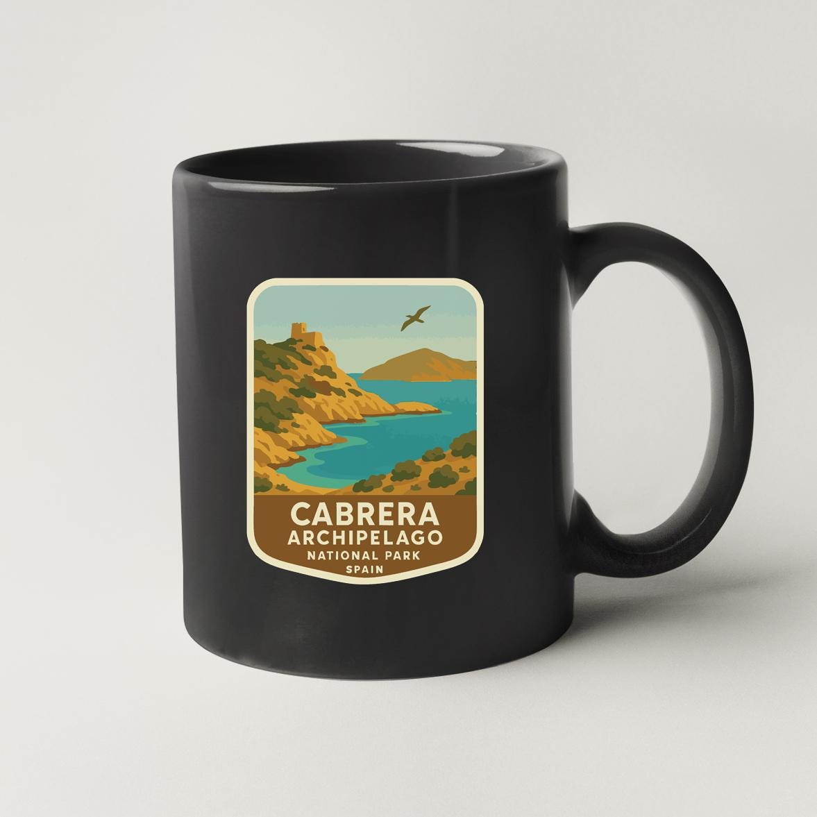A black coffee mug with a label that features an illustration of a coastal landscape with mountains, a bird flying overhead, and a body of water.