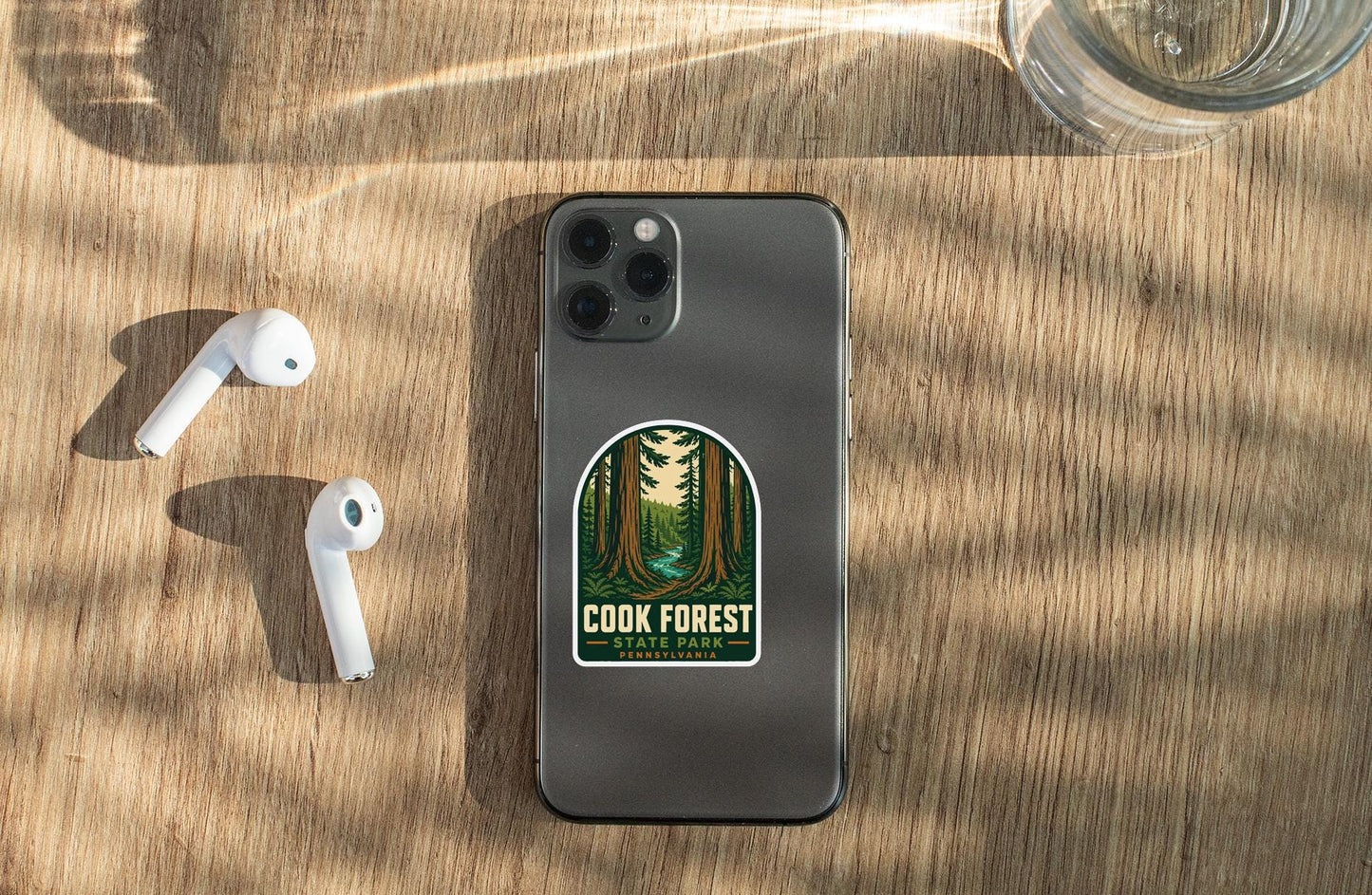 a smartphone with a sticker on the back, placed on a wooden surface alongside a pair of white earbuds and a glass of water.