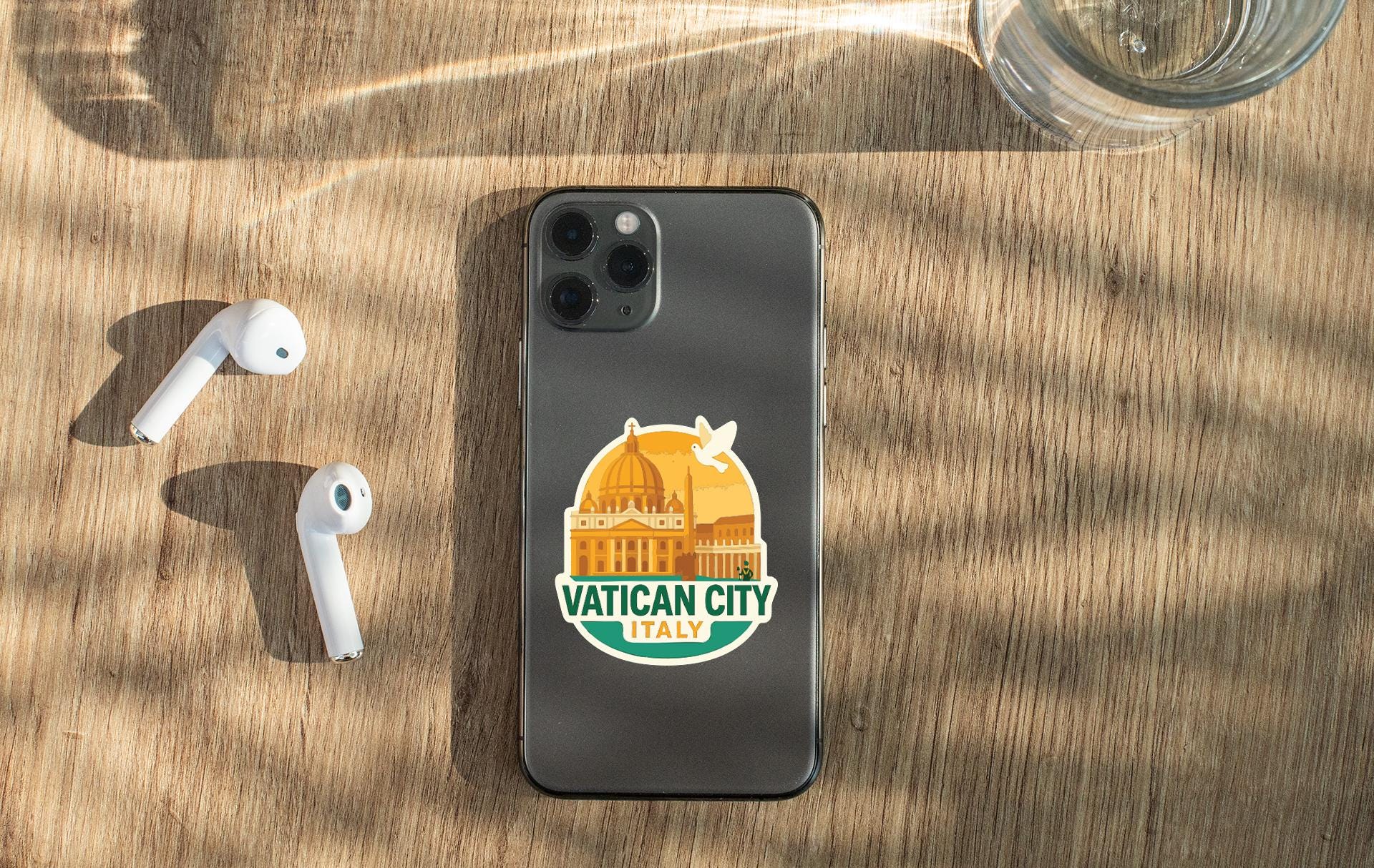 a smartphone with a sticker on the back, placed on a wooden surface alongside a pair of white earbuds and a glass of water.