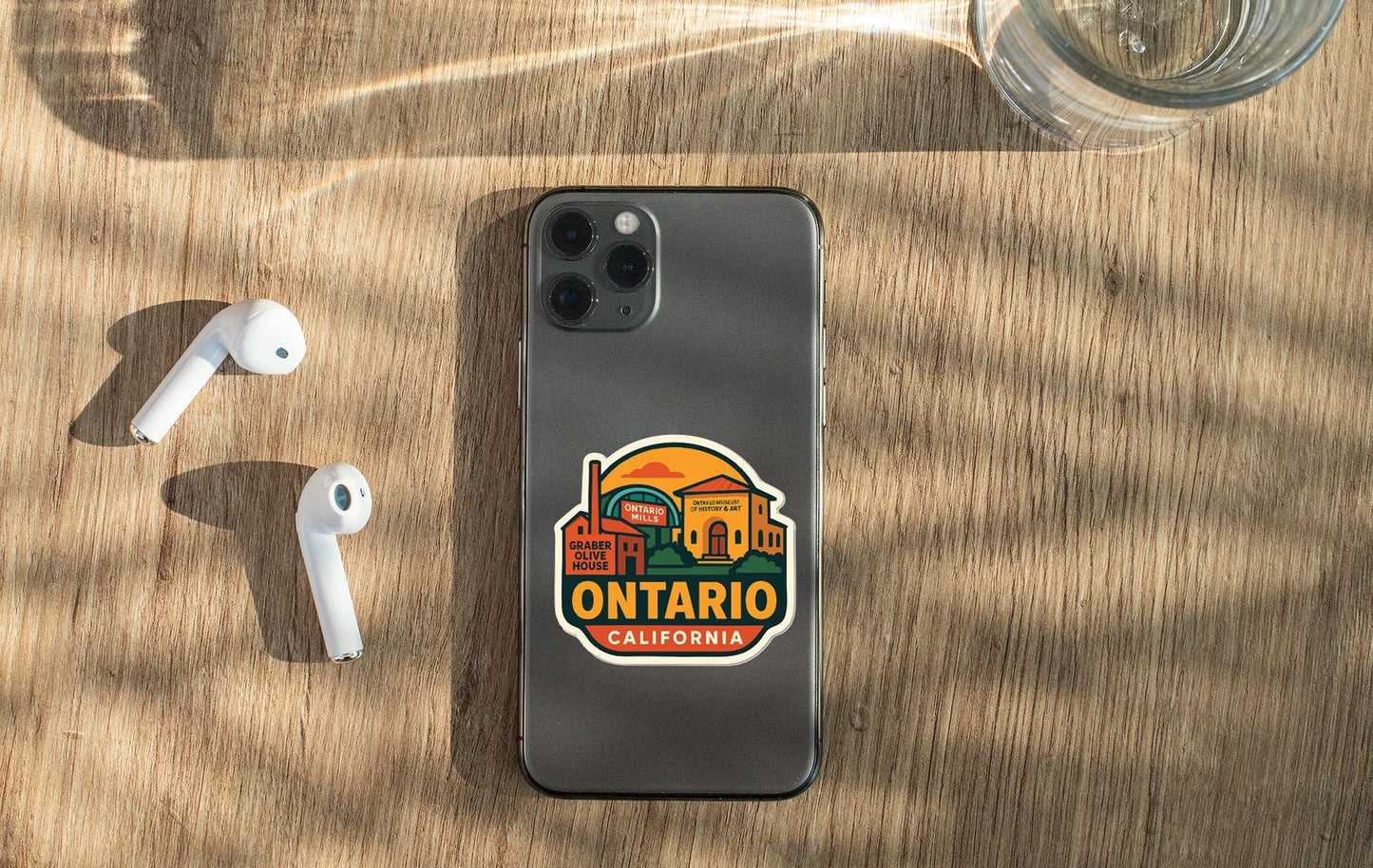 a smartphone with a sticker on the back, placed on a wooden surface alongside a pair of white earbuds and a glass of water.
