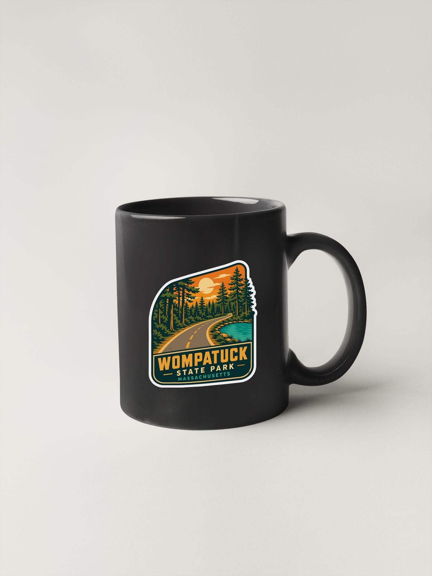 A black coffee mug with a sticker depicting a scenic road leading through a forest, with the text "WOMPATUCK STATE PARK MASSACHUSETTS" printed on it.