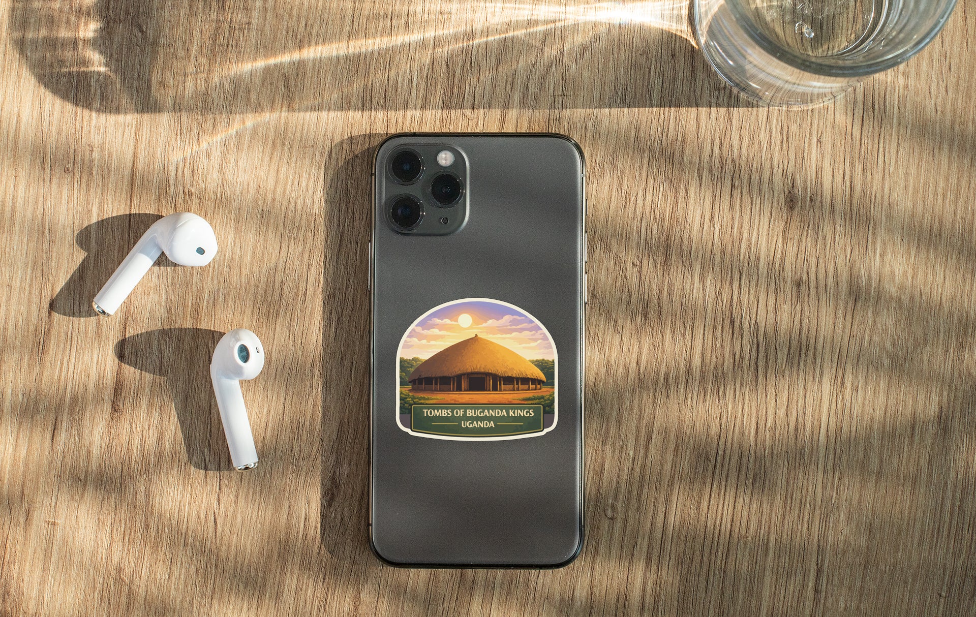 a smartphone with a sticker on the back, placed on a wooden surface alongside a pair of white earbuds and a glass of water.