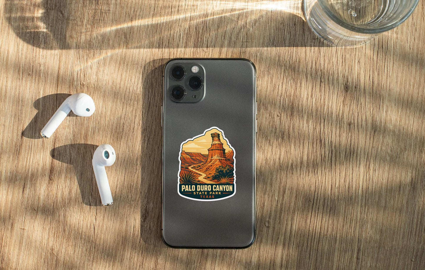 a smartphone with a sticker of the Palid Duro Canyon State Park on it, along with a pair of white earbuds and a glass of water on a wooden surface.