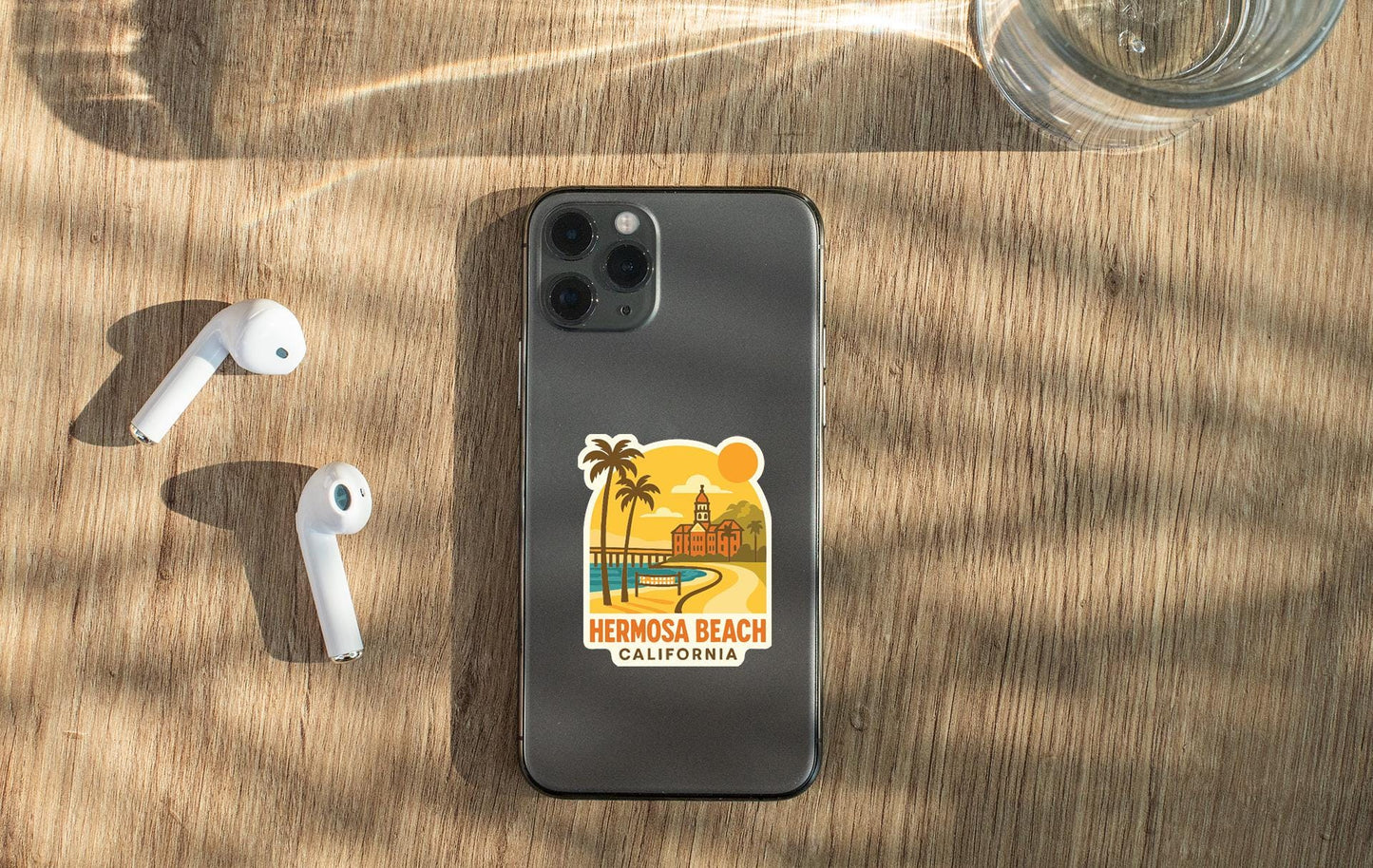 a smartphone with a sticker of Hermosa Beach, California, placed on a wooden surface alongside a pair of white earbuds and a glass of water.
