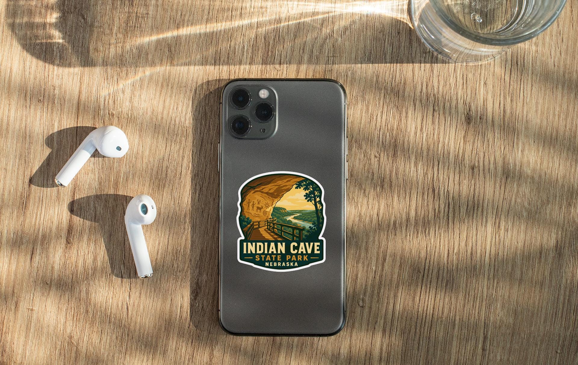 a smartphone with a sticker on it, placed on a wooden surface alongside a pair of white earbuds and a glass of water.