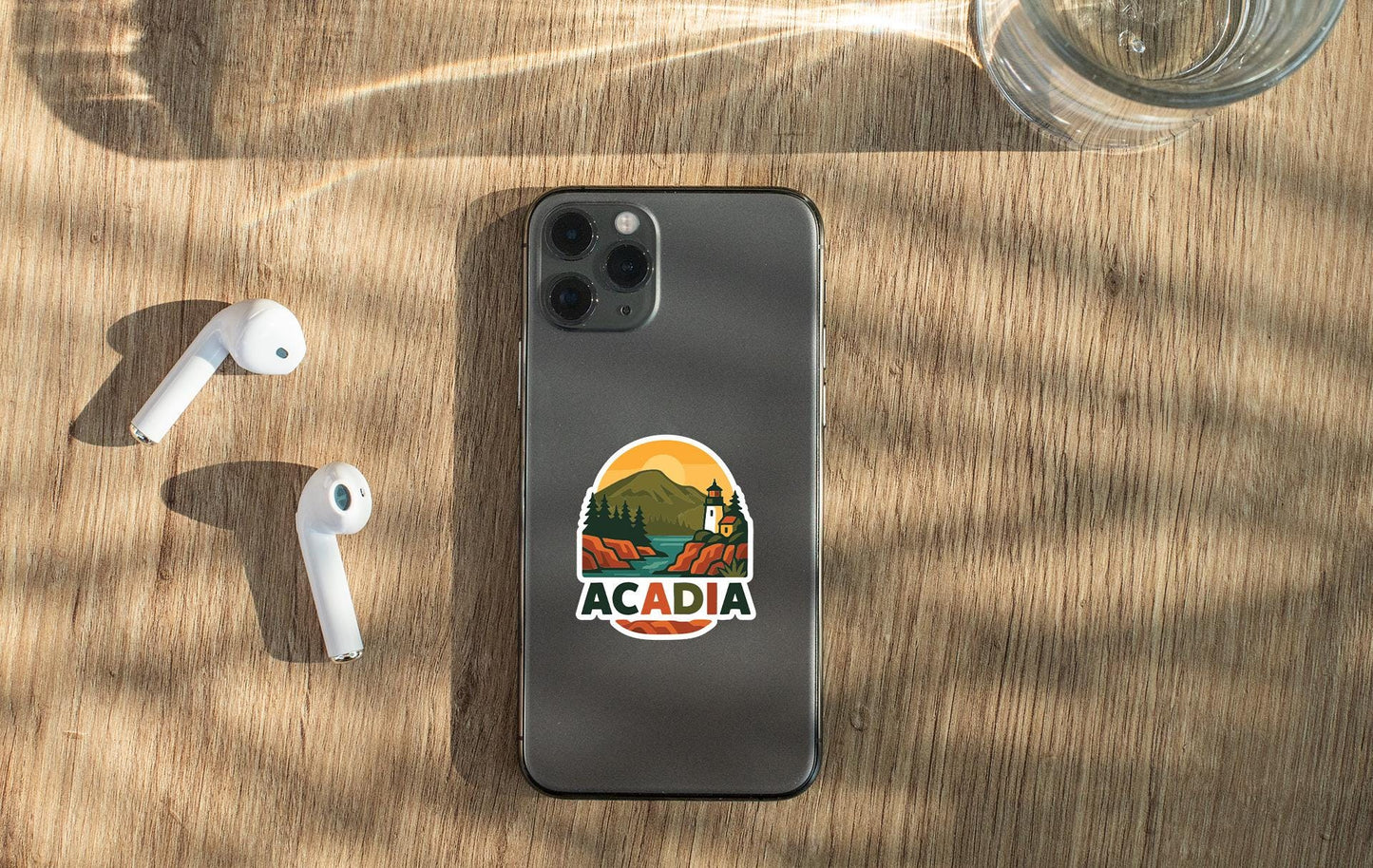 a smartphone with a sticker of Acadia National Park on its back, along with a pair of white earbuds and a glass of water on a wooden surface.
