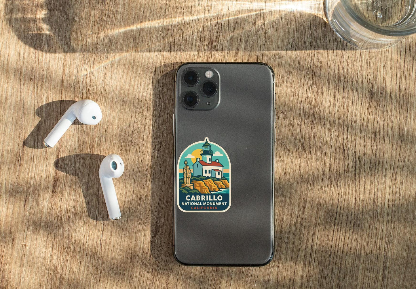 a smartphone with a sticker on it, placed on a wooden surface alongside a pair of white earbuds and a glass of water.