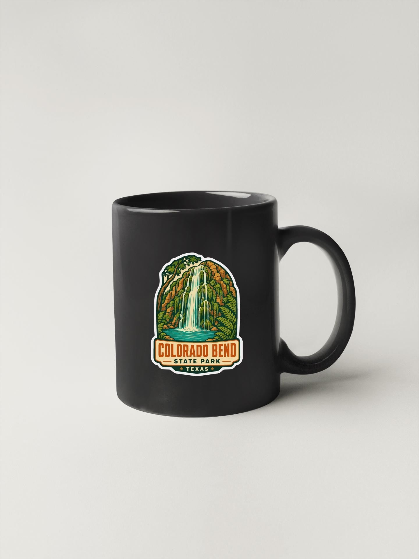 A black coffee mug with a colorful graphic depicting a waterfall and the text "COLORADO BEND STATE PARK" printed on it.
