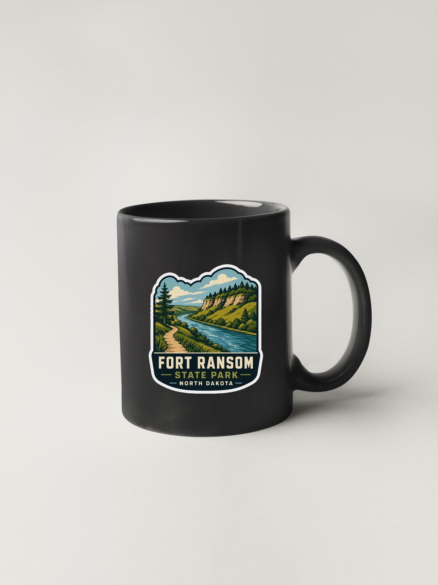 A black coffee mug with a graphic design depicting a scenic landscape, including a river, mountains, and trees.
