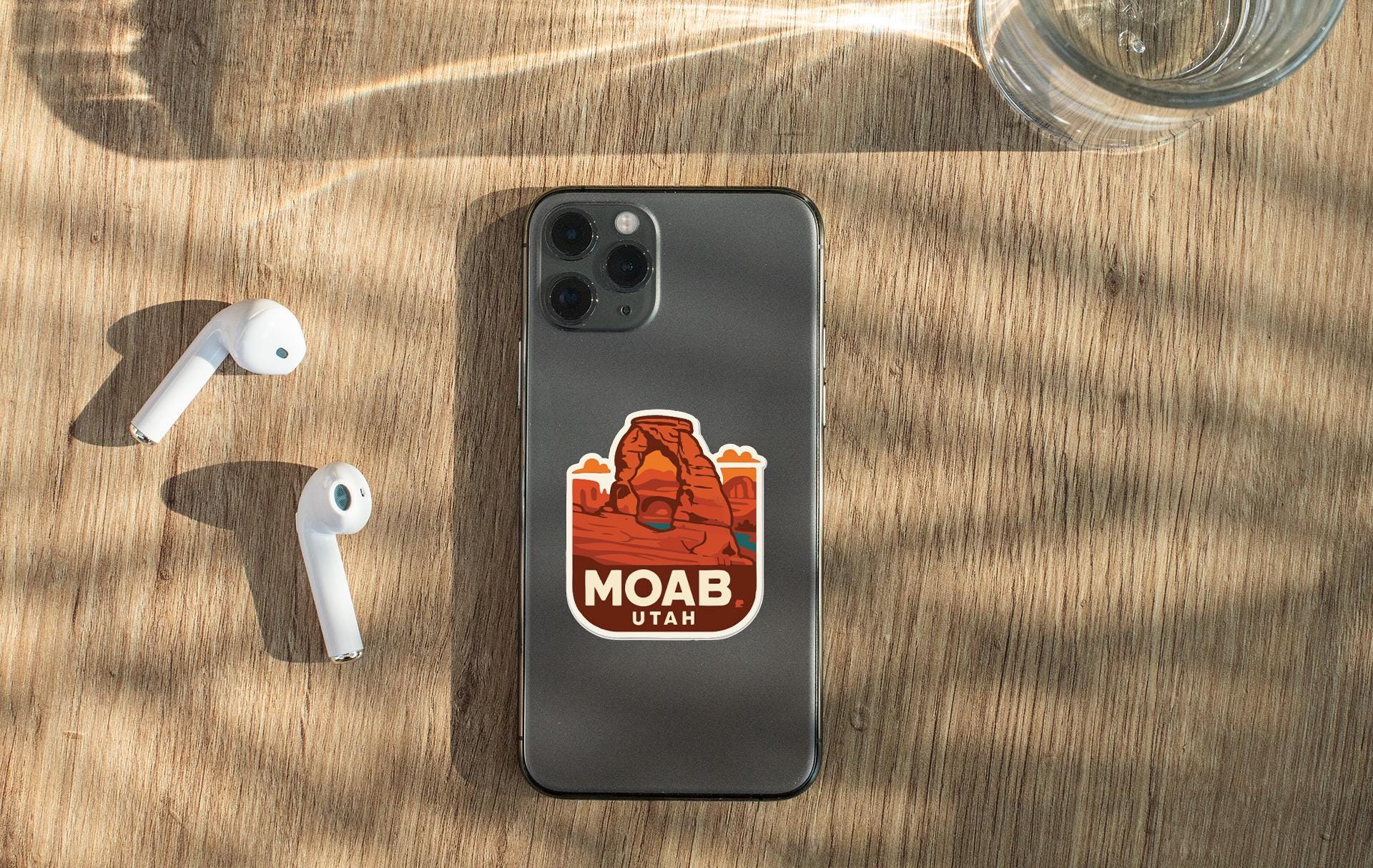 a smartphone with a sticker of Moab Utah on it, placed on a wooden surface alongside a pair of white earbuds and a glass of water.