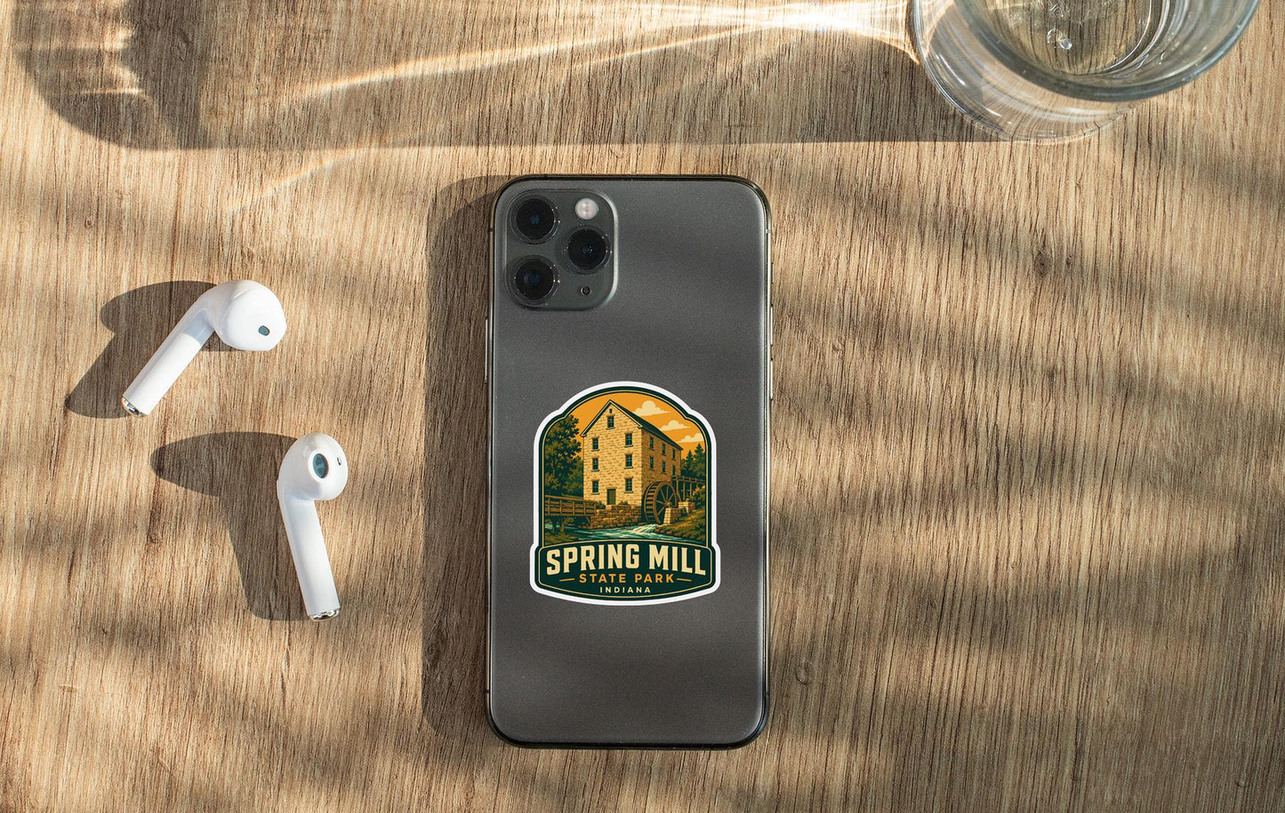 a smartphone with a sticker on it, placed on a wooden surface alongside a pair of white earbuds and a glass of water.