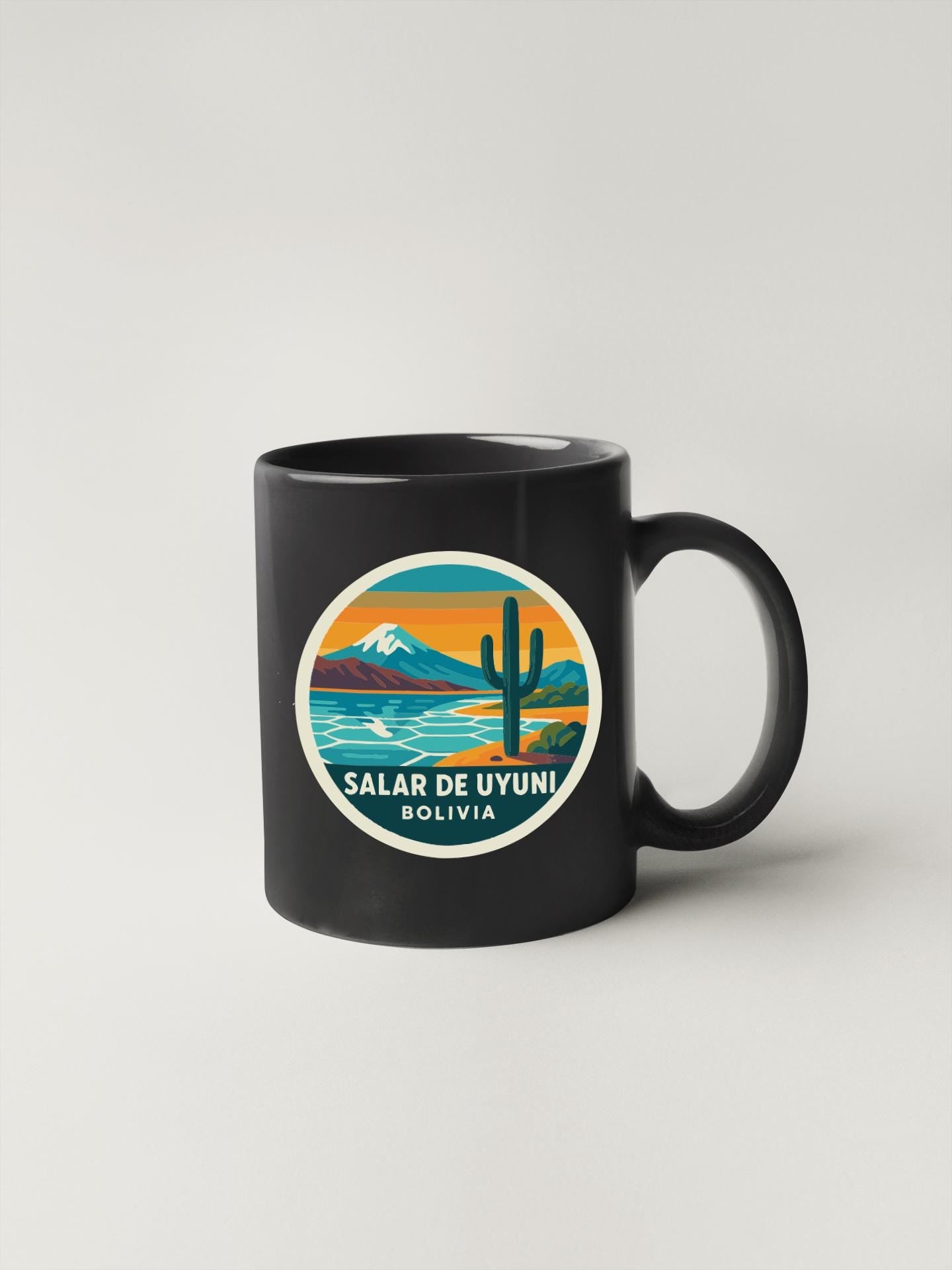 A black coffee mug with a circular design featuring a desert landscape with a cactus, mountains, and a body of water.